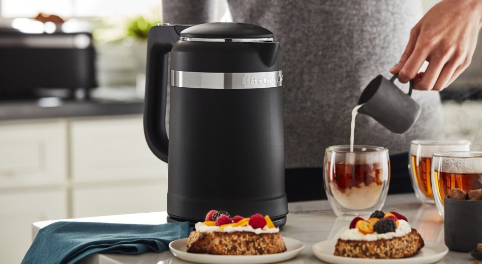 Hand pouring milk into cups of tea sitting next to electric tea kettle and plates of pastries.